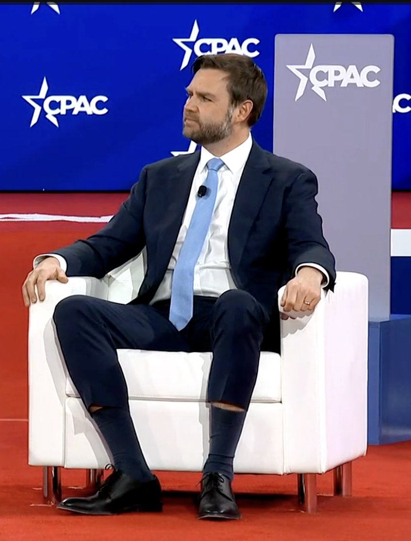 JD Vance sits in a white chair on the CPAC stage. He is wearing a light blue tie that waterfalls down over his crotch, and skinny-leg suit pants. Sitting down has made the pantlegs hike up o