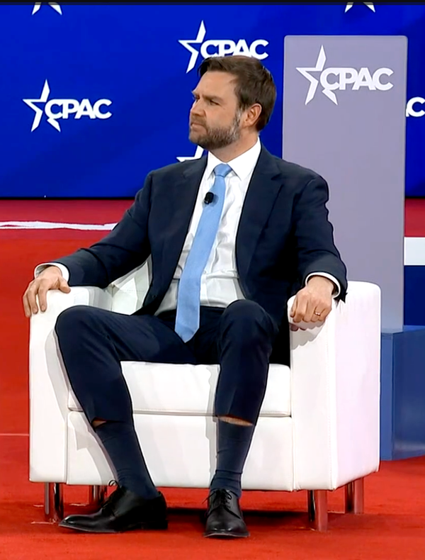 JD Vance sits in a white chair on the CPAC stage. He is wearing a light blue tie that waterfalls down over his crotch, and skinny-leg suit pants. Sitting down has made the pantlegs hike up o